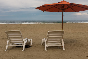 Two empty plastic white chairs under an orange beach umbrella on a deserted sandy beach in Costa Rica