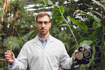 handsome scientist in white coat and glasses holding rubber gas mask and flask with plant sample in orangery