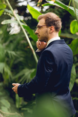 businessman in suit and glasses talking on smartphone in greenhouse and looking away