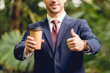 cropped view of smiling businessman in suit and tie holding coffee to go and showing thumb up in orangery