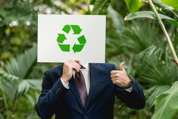 businessman holding white card with green recycling sign in front of face and showing thumb up in...