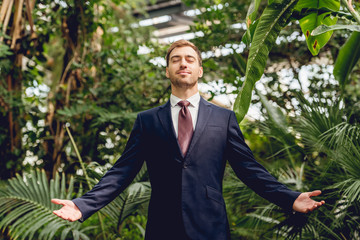 joyful businessman with closed eyes and outstretched hands standing in greenhouse