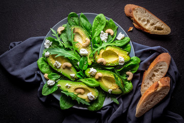 Boneless avocado slices with arugula and spinach salad sprinkled with rehas cashews and sesame seeds on a gray plate and gray linen fabric with baguette pieces on a black background. top view