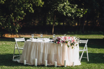 A large white round table, decorated with fresh flowers, candles stands on the nature, in a park with green grass. Wedding decorations and details. Preparing for a wedding party.
