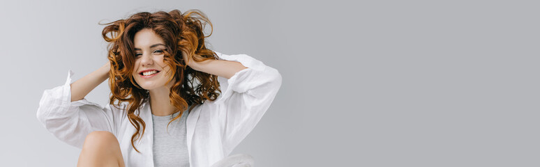 panoramic shot of cheerful girl touching red curly hair and smiling on grey