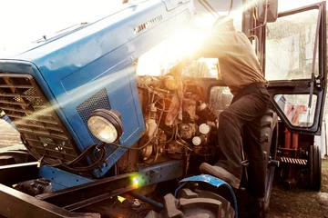 Crédence de cuisine en verre imprimé Tracteur Mécanicien agriculteur réparant le moteur du tracteur bleu. Réparer la technologie agricole au coucher du soleil.