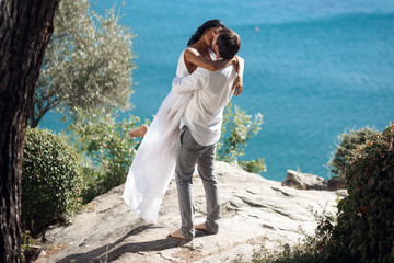 Man holding woman in his arms wrapped around her waist and she is holding on to him. The wind is blowing her hair across her face, standing in a seascape near the mediterranea sea in Greece.