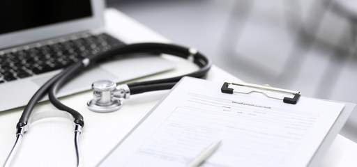 Stethoscope, clipboard with medical form lying on hospital reception desk with laptop computer and busy doctor and patient communicating at the background. Medical tools at doctor working table