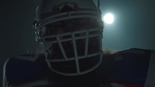 Close-up portrait of american football player in sports equipment and helmet pointing his finger in camera on dark background with spotlight. Front view