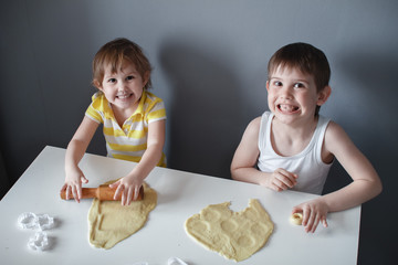 Little cheerful children make shortbread. Rolls out the dough with a rolling pin on a white table. The preparation of the dessert