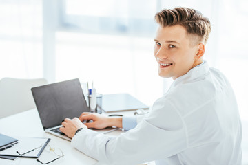 cheerful doctor smiling while using laptop with blank screen