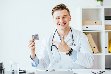 handsome and cheerful doctor in white coat pointing with finger at condom