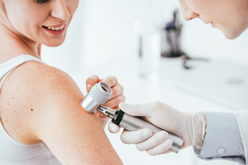 cropped view of dermatologist holding dermatoscope while examining cheerful woman with skin disease - obrazy, fototapety, plakaty