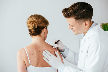 happy dermatologist in latex gloves holding marker pen near woman with melanoma isolated on white
