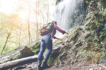 young woman with backpack hiking near waterfall in forest 