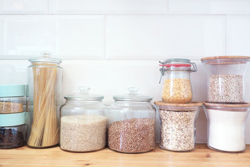 Glass jars with products stand on a wooden tabletop against the background of white ceramic tiles in the form of bricks. ..