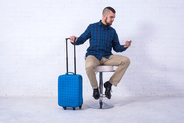 A man with a blue suitcase and passport is sitting on a chair near a white brick wall.