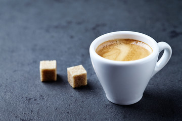 Cup of coffee and two brown sugar cubes on dark stone background. Close up. 