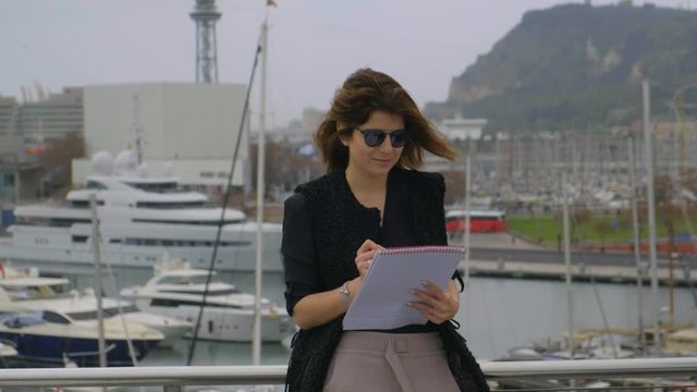 Beautiful woman writing to the notebook while wind touching her hair on the rooftop at the port with yachts 