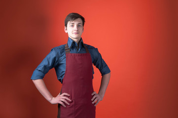 handsome barista with dark hair in burgundy apron looking at camera and holding hands on hips on...
