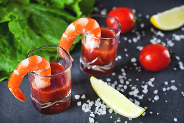 Shrimp cocktail in small glasses, with cherry tomatoes, lemon with salt and spices, salad leaves with shrimps, side view on a dark gray background