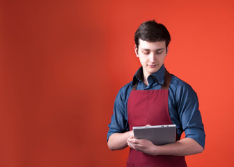 handsome barista with dark hair in burgundy apron holding and looking at digital tablet on orange background with copy space