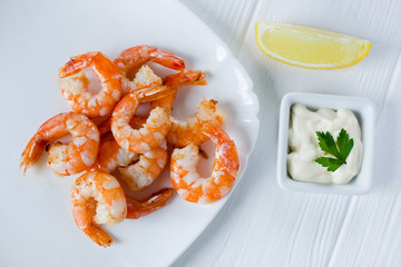 Fried peeled shrimps in a white plate on a wooden white table with white sauce and lemon, top view