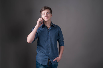 confident handsome young man in blue shirt talking at smartphone, smiling and looking away on grey background with copy space