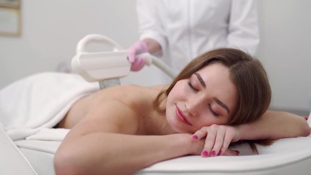 Young pretty smiling woman back laser epilation in beauty salon. Cosmetologist doing depilation treatment with elos device. Beautician hands in gloves holding hair removal cosmetology ipl hardware