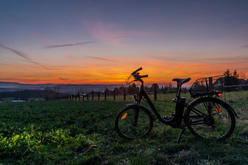 Naklejka na ściany i meble Black electric bicycle with sunset on light green meadow in spring evening