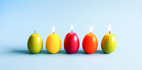 Bright burning paraffin candles in the shape of colorful Easter eggs on blue background.