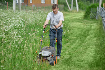 Gardener is working in the yard of a countryside cottage, he is using lawn mower.