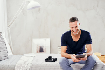 Caucasian man in forties sitting on bed in bedroom and using tablet.