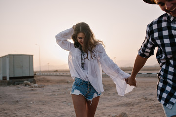 Young loving laughing couple in trendy clothes running on sand holding hands at sunset. Smiling...