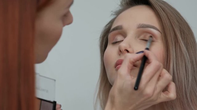 Makeup artist makes makeup beautiful blonde girl on a light monophonic background. Paints eyes with mascara, applies powder with a brush. Makeup artist studio concept.