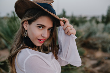 Elegant long-haired girl in stylish hat looks back, while walking in beautiful exotic park. Close-up portrait of pretty young woman in trendy earrings and shirt posing with enigmatic face expression