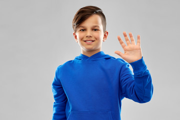 childhood, gesture and people concept - portrait of smiling boy in blue hoodie waving hand over grey background © Syda Productions
