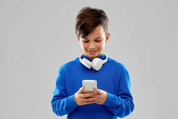 childhood, technology and people concept - smiling boy in blue hoodie with headphones on neck using smartphone over grey background