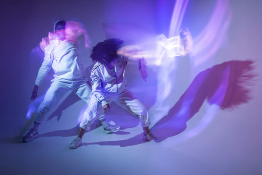 Young man and woman in sportswear performing energetic dance movements and leaving light traces under bright violet illumination