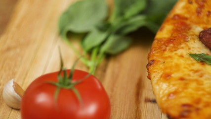 Fresh hot appetizing peperoni pizza lying on a wooden plate on the table decorated with basil greens, tomato and garlic in slow motion close up front view 4K video