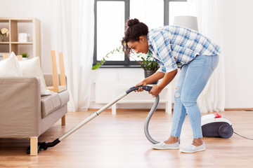 household and housework concept - happy african american woman or housewife with vacuum cleaner cleaning floor under sofa at home - obrazy, fototapety, plakaty
