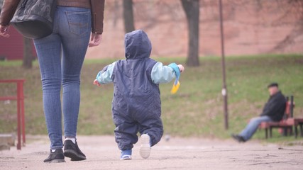 Young woman with her small son who holding baby shovel are walking near a pond in the city park in a spring cloudy day in 4K back view slow motion video.
