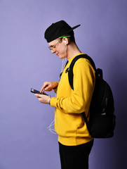 Portrait of a cheerful student wearing backpack, in cap and glasses and using smartphone over purple background