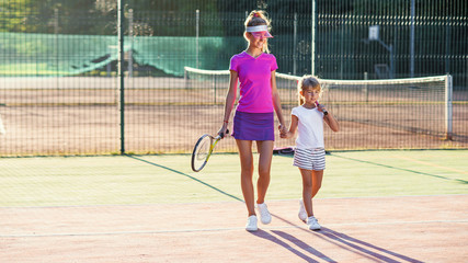 Obraz premium Female tennis trainer holds her little student to the hands and they goes together after training at outdoor tennis court at sunset.