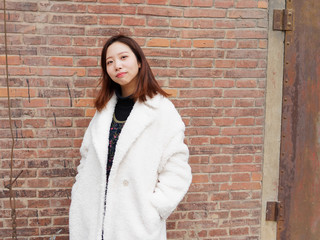 Street photography of a beautiful Chinese woman in white coat with red brick wall background, looking at you with two hands in her pocket, portrait of cute girl with nevus near eyes.