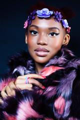 young pretty african american woman in spotted fur coat and flowers jewelry on black background close up