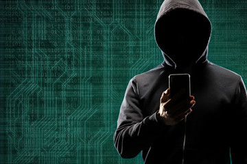 Dangerous hacker with a smartphone gadget over digital background with binary code. Obscured dark face in mask and hood. Data thief, internet attack, darknet fraud, virtual reality and cyber security.
