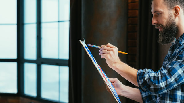 Modern loft studio. Side view portrait of young handsome enthusiastic lefthanded artist standing with sketch board and brush.