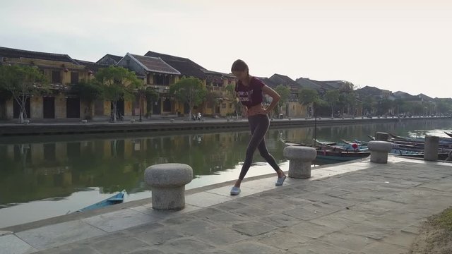 pictorial picture beautiful girl trains doing squats on paved embankment near river against yellow buildings in morning
