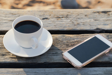 White Cup on black coffee  and smartphone on beach at sunrise or sunset for lifestyle and blogger concept. Remote work. Copy space.